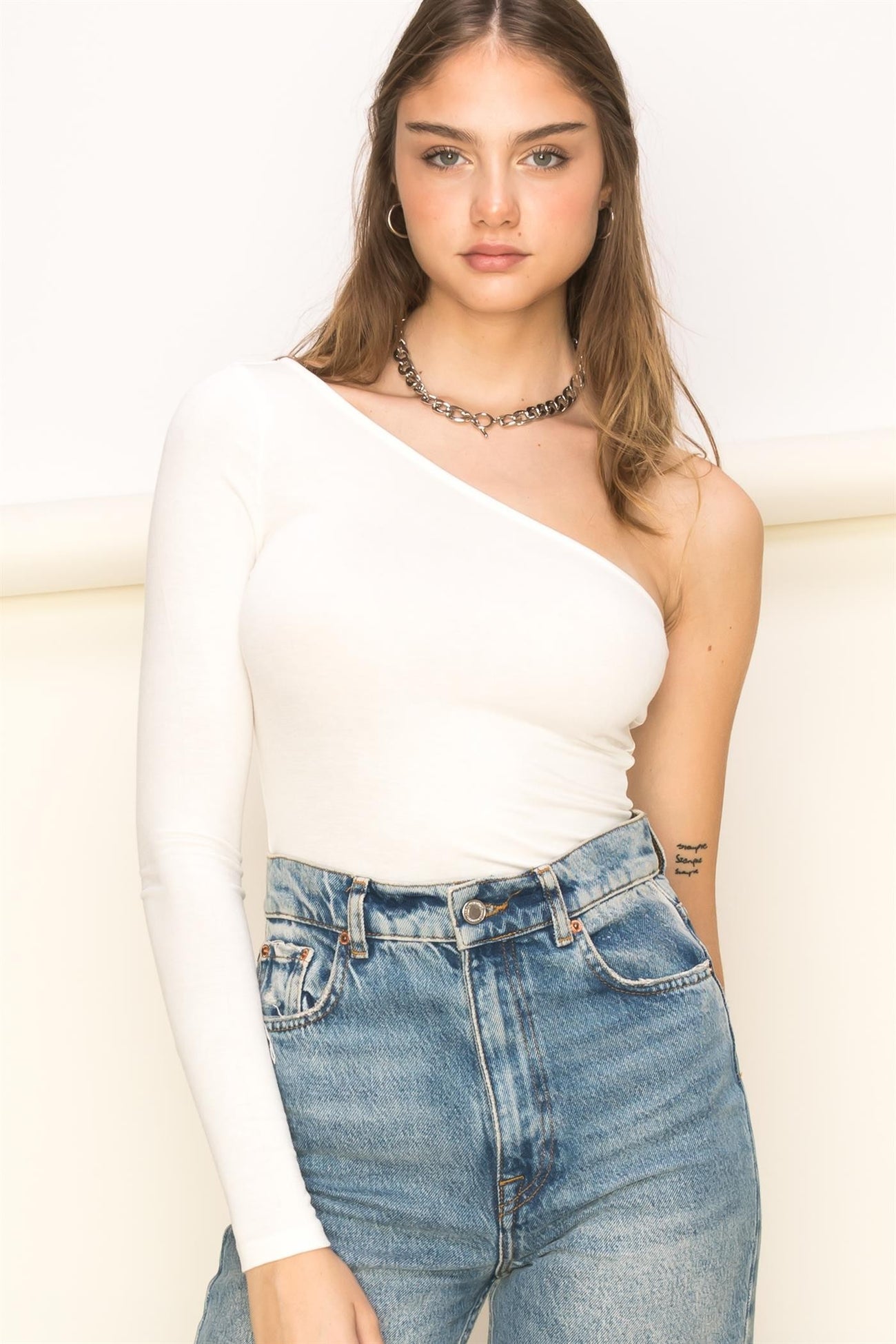 woman wearing a white one-shoulder fitted top with a long sleeve, paired with high-waisted denim jeans