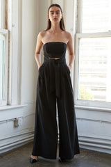 woman wearing a black strapless jumpsuit with a tied waist and wide-leg pants, standing against a bright window