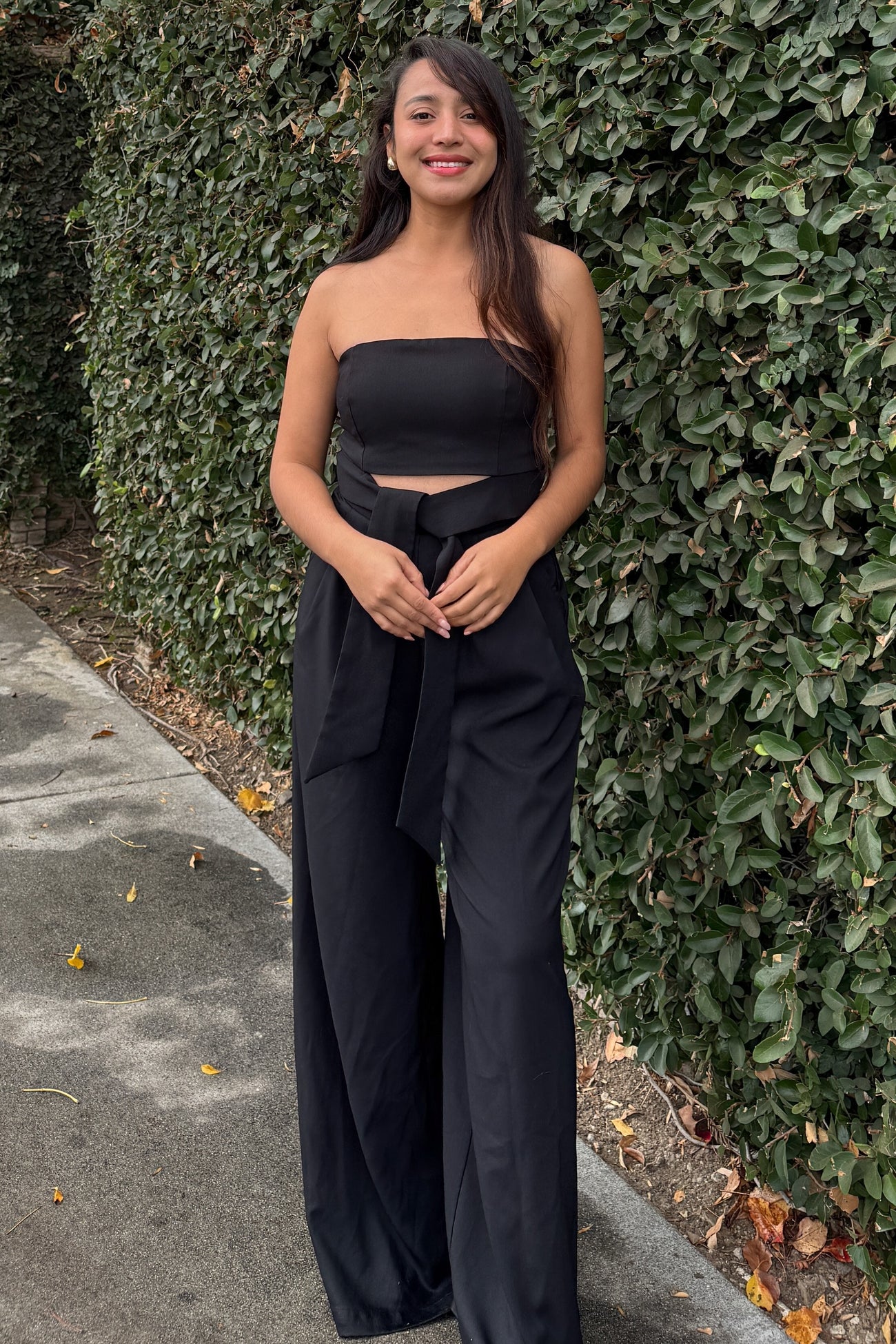 woman wearing a black strapless jumpsuit with a cutout detail and a tied waist, standing outdoors against greenery