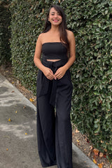 woman wearing a black strapless jumpsuit with a cutout detail and a tied waist, standing outdoors against greenery