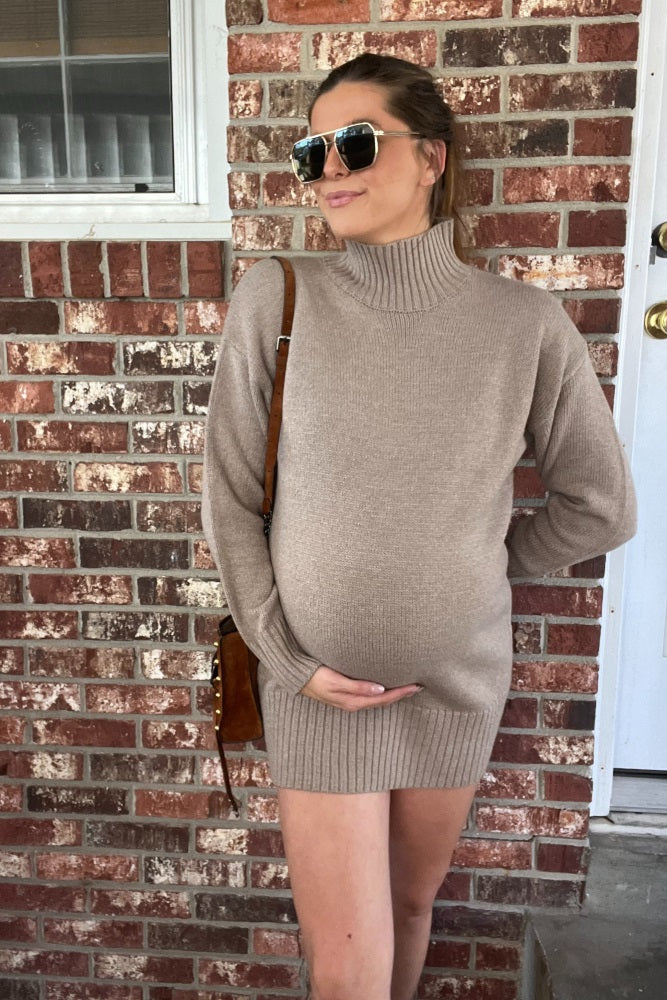 woman wearing a beige maternity dress with a turtleneck, holding her baby bump against a brick wall