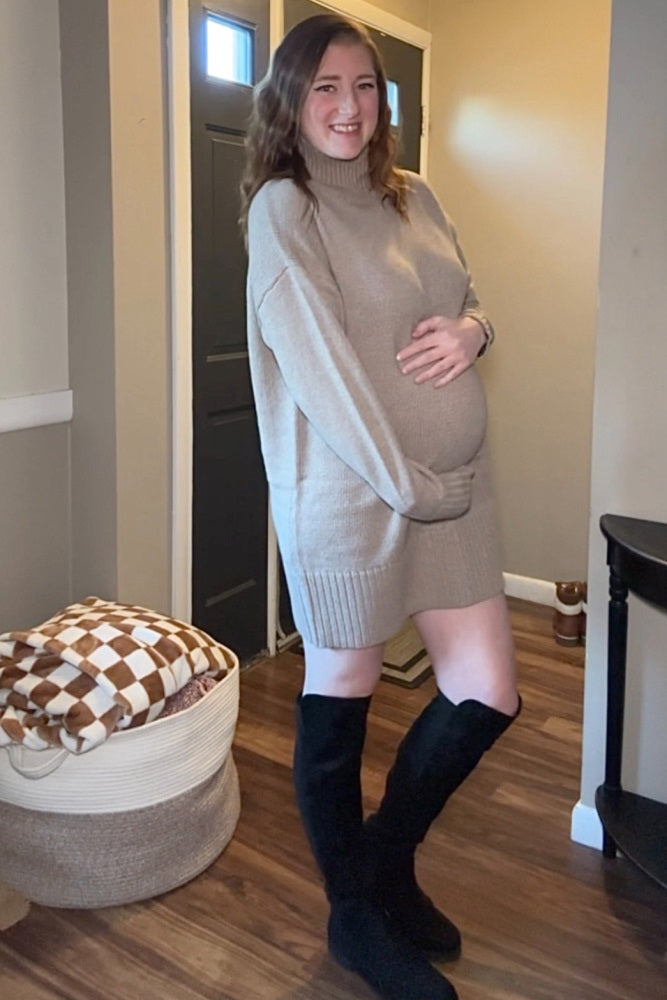 woman wearing a beige oversized maternity sweater dress and showing her baby bump while standing indoors
