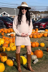 woman wearing a beige maternity sweater dress and brown knee-high boots, holding her baby bump in a pumpkin patch