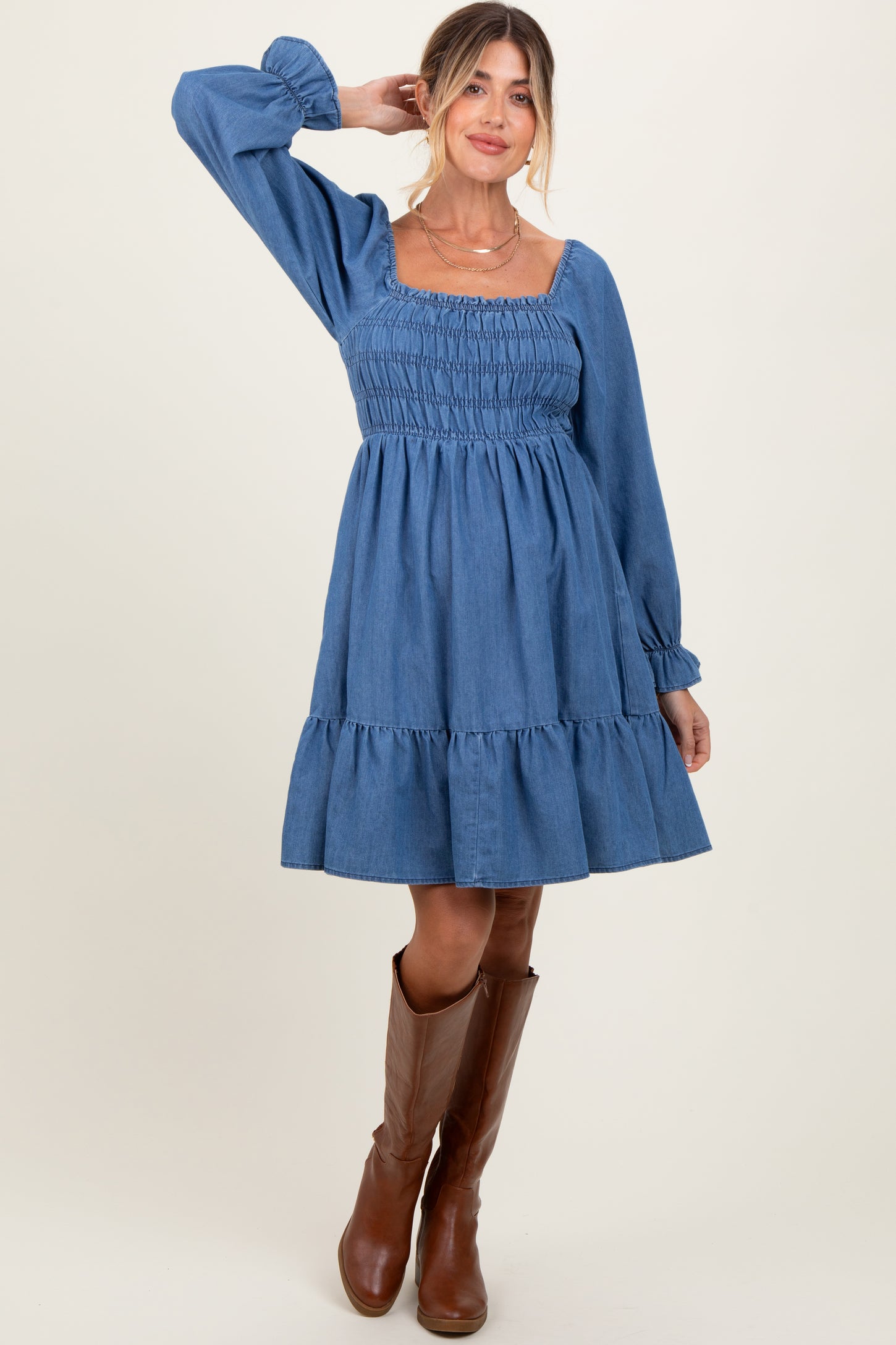 woman wearing a denim maternity dress with smocked bodice and ruffled hem, paired with brown knee-high boots