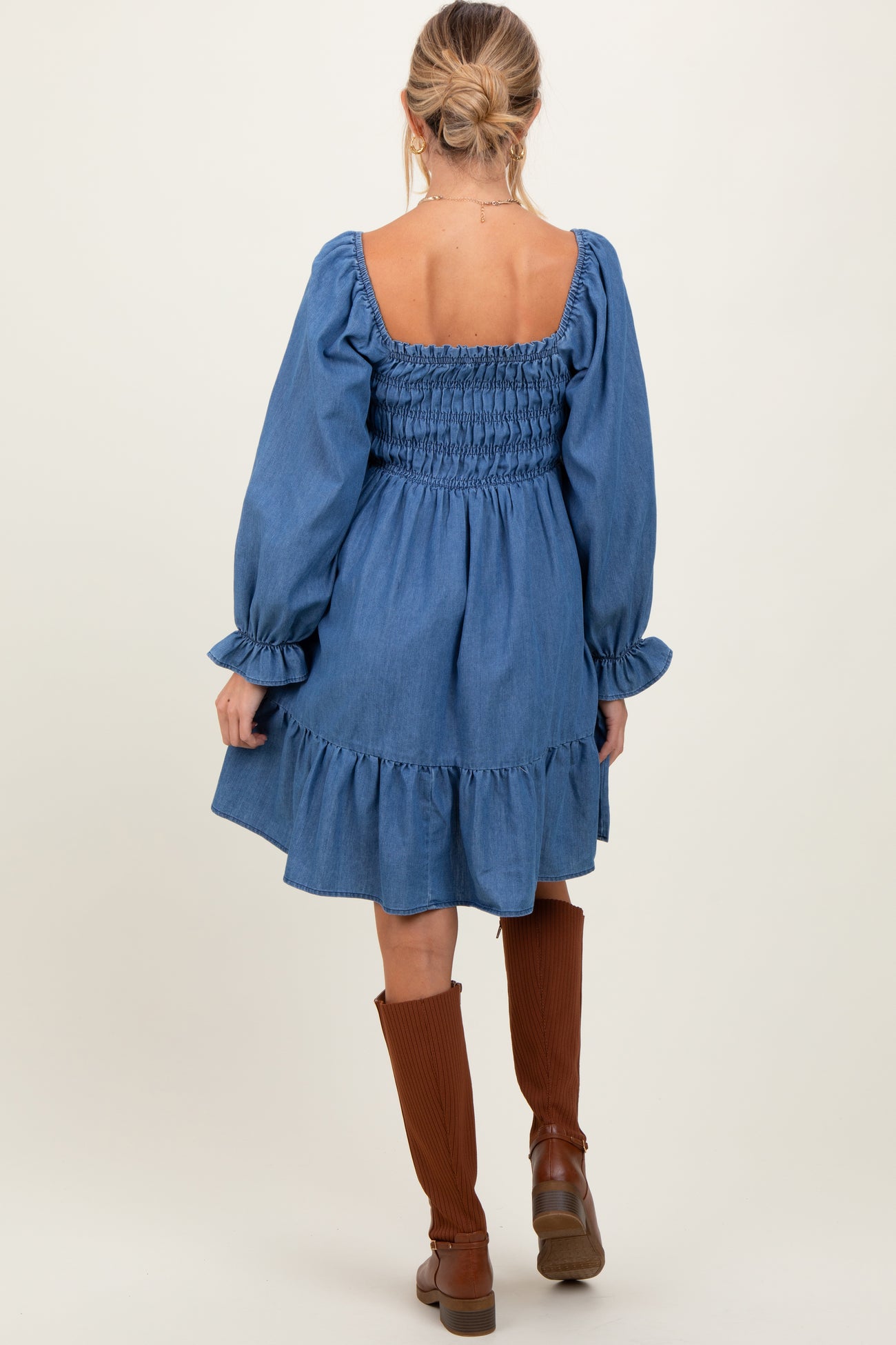 woman wearing a blue denim maternity dress with a smocked back and ruffled hem, paired with brown knee-high boots
