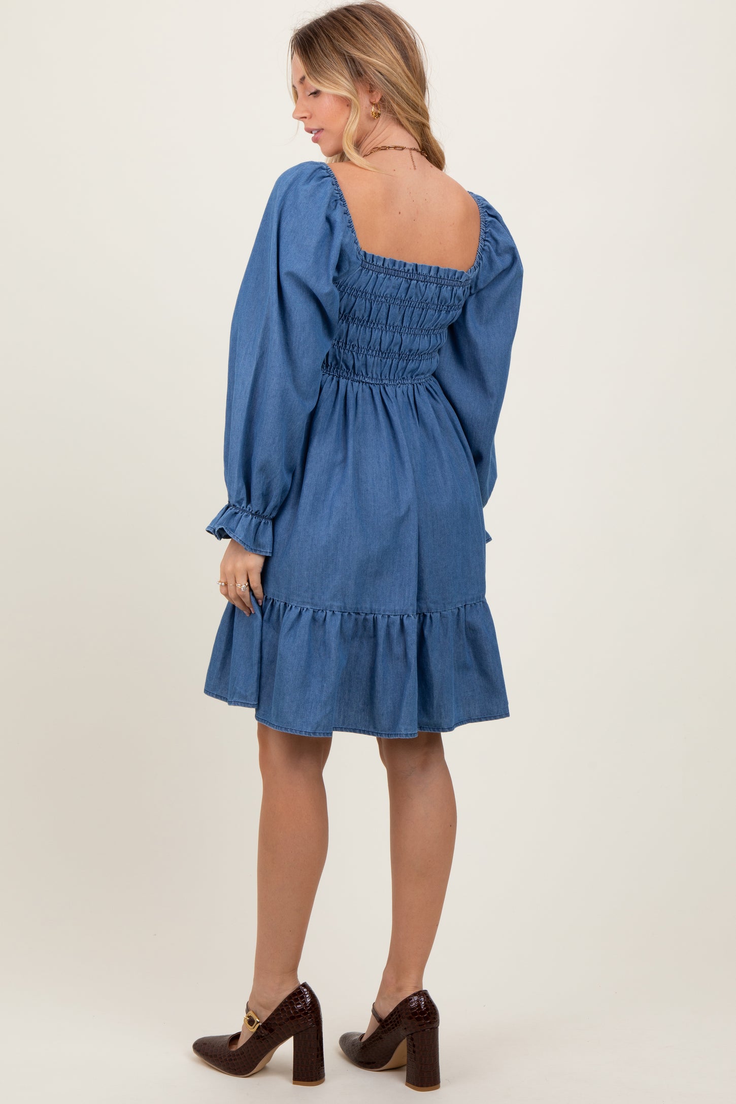 woman wearing a denim smocked dress with long sleeves and a ruffled hem, showing the back view