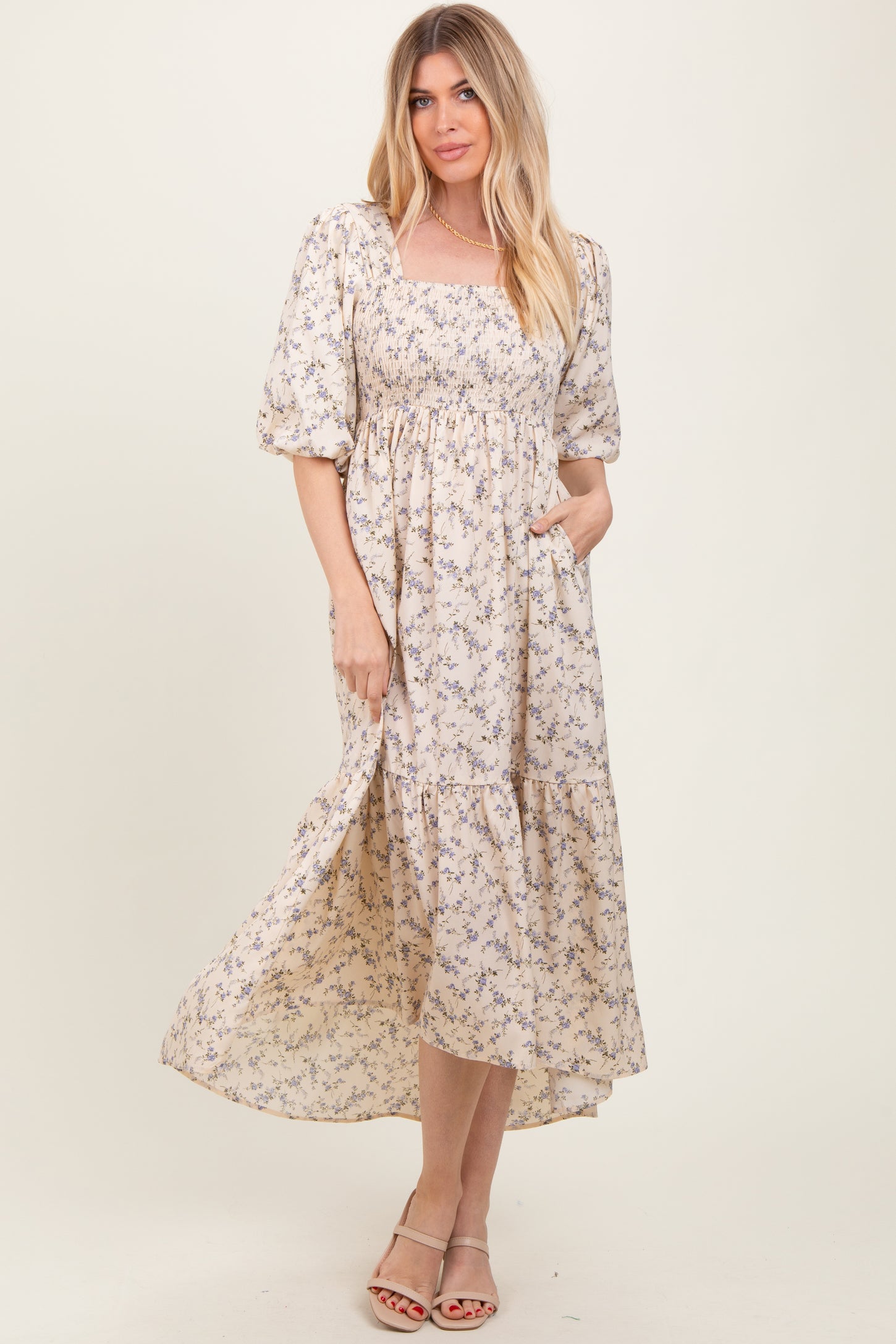 woman wearing a beige floral maxi dress with puff sleeves and a smocked bodice, standing with one hand in the pocket