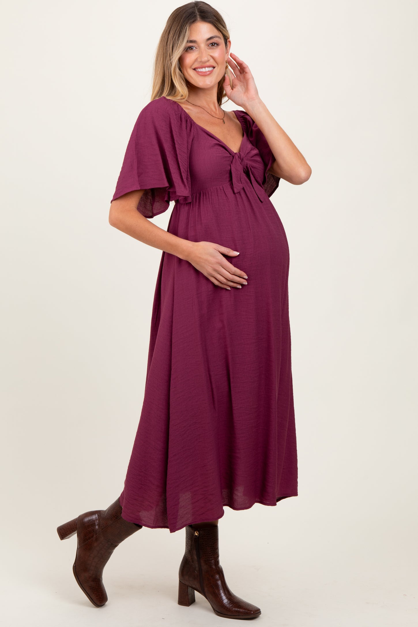 woman wearing a burgundy maternity dress with a tie front and flutter sleeves, gently cradling her baby bump