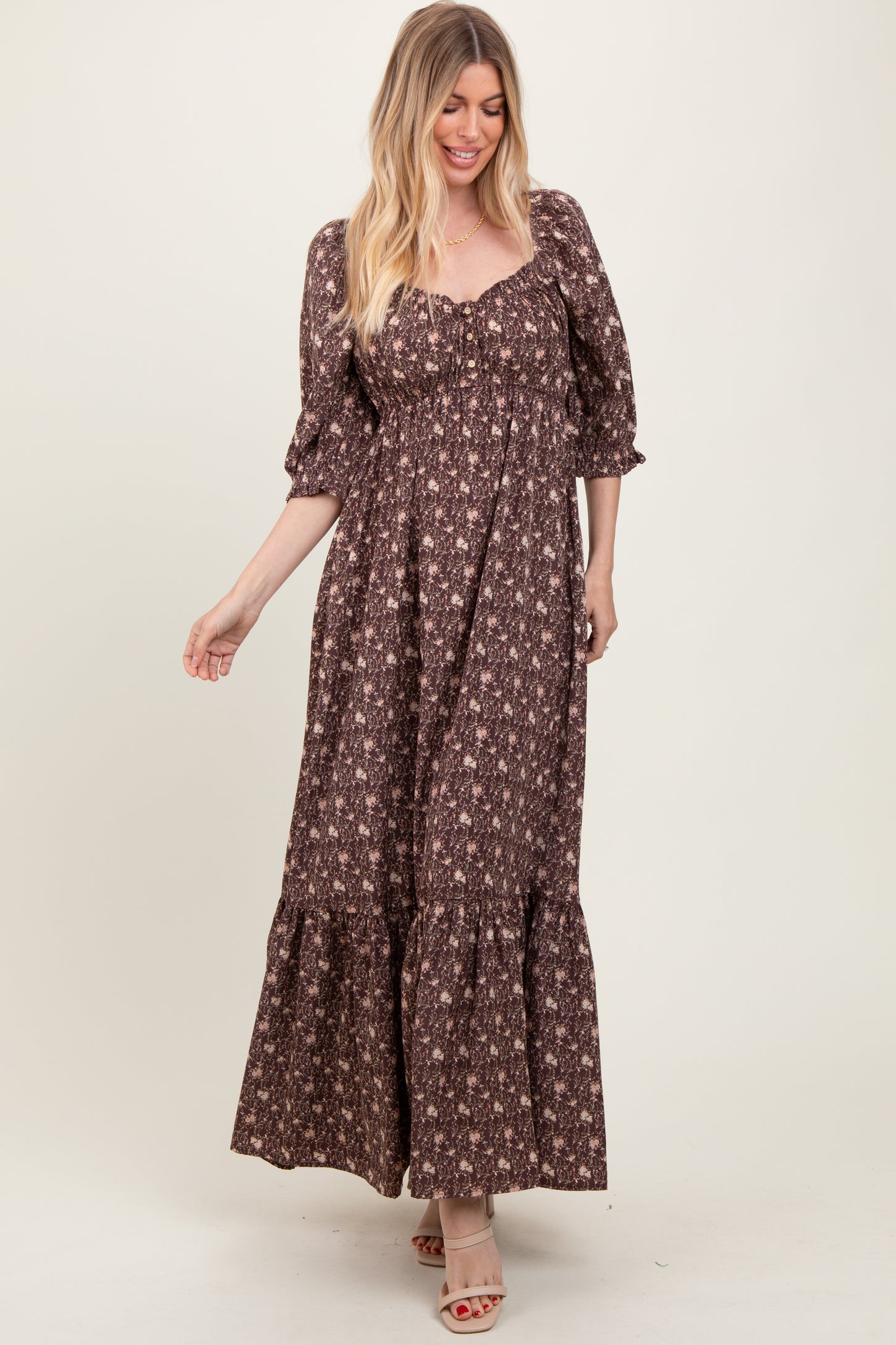 woman wearing a brown floral maxi dress with puff sleeves and a tiered hem, standing and smiling