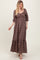 woman wearing a brown floral maxi dress with puff sleeves and a tiered hem, standing and smiling