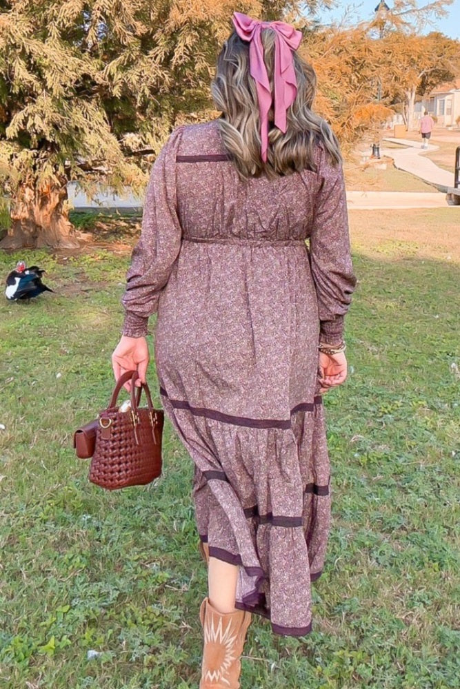 woman wearing a mauve floral maxi dress with tiered detailing and a ribbon in her hair, carrying a small brown handbag