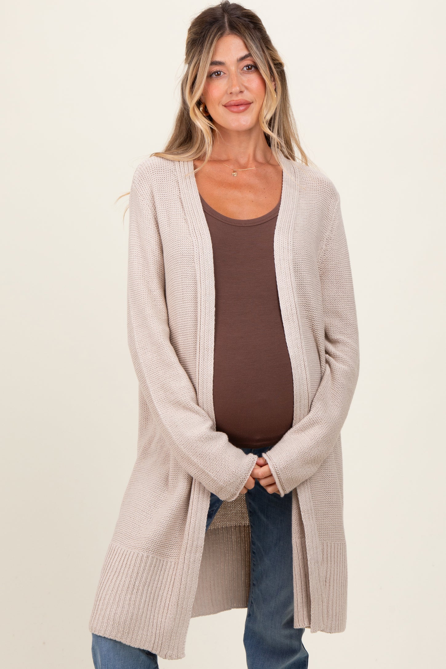 woman wearing a beige maternity cardigan over a brown ribbed tank top and holding her baby bump