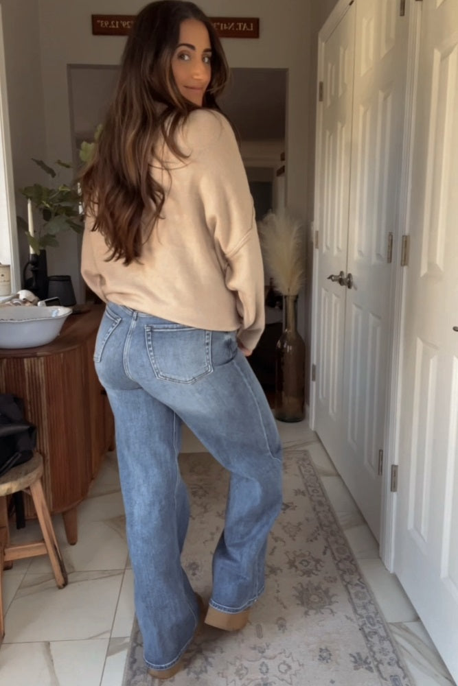 woman wearing a tan oversized sweatshirt and light blue wide-leg jeans, standing in a stylish indoor setting