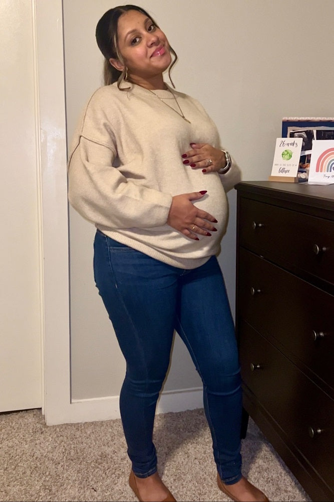 woman wearing a beige oversized sweater and blue maternity jeans, holding her baby bump