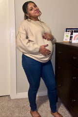 woman wearing a beige oversized sweater and blue maternity jeans, holding her baby bump