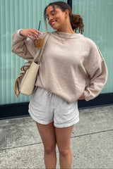 woman wearing a light beige oversized sweater and white drawstring shorts, holding a drink and smiling