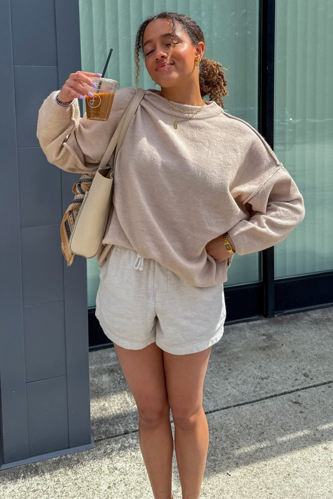 woman wearing a beige oversized sweater and light beige shorts, holding an iced coffee and a handbag