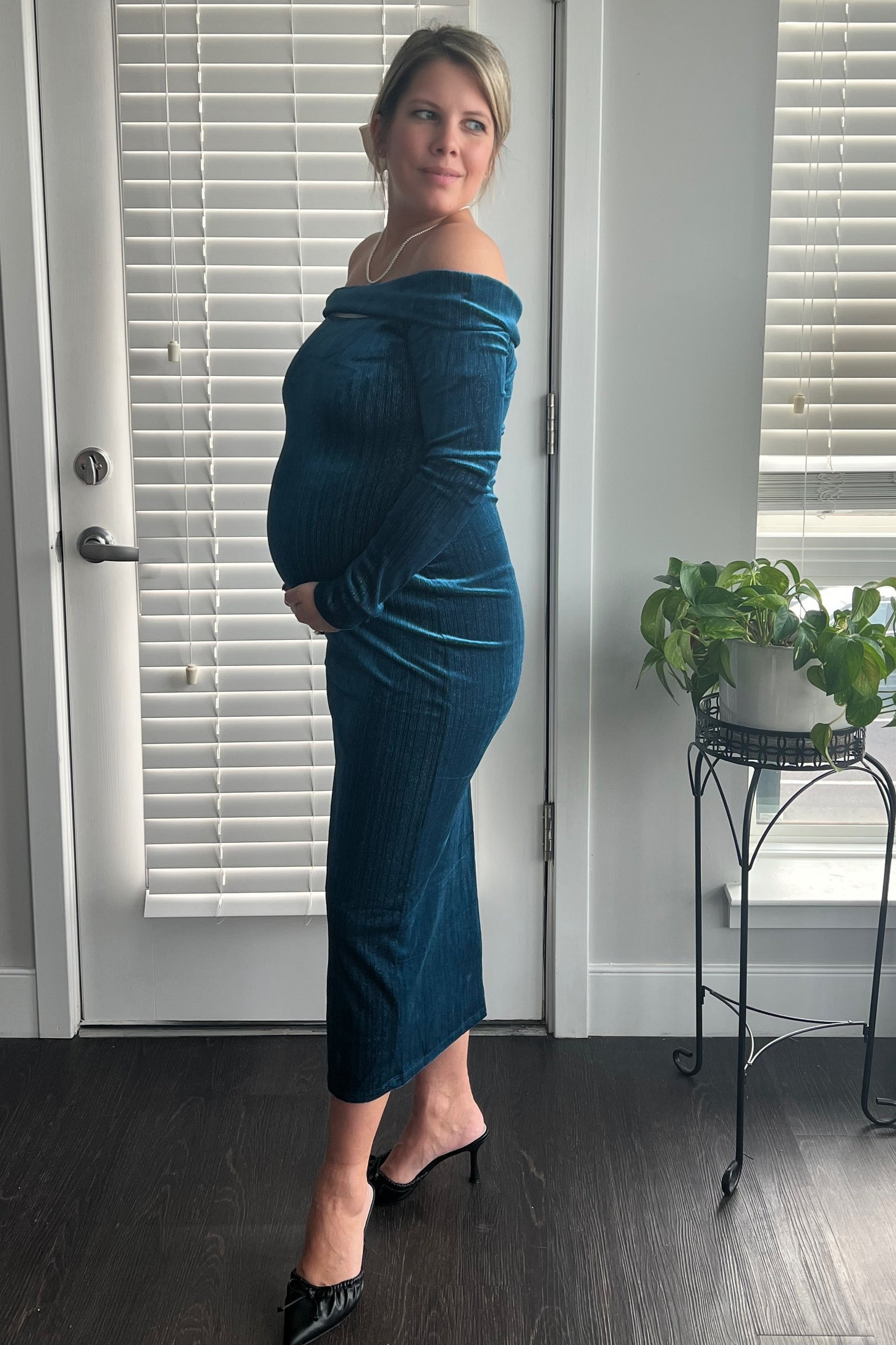 woman wearing a dark teal off-shoulder maternity dress with a fitted silhouette and holding her baby bump