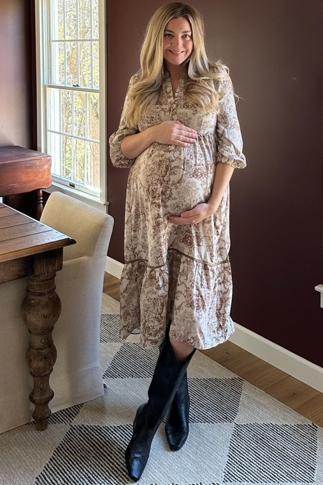 woman wearing a floral beige maternity dress and holding her baby bump while standing in a bright room