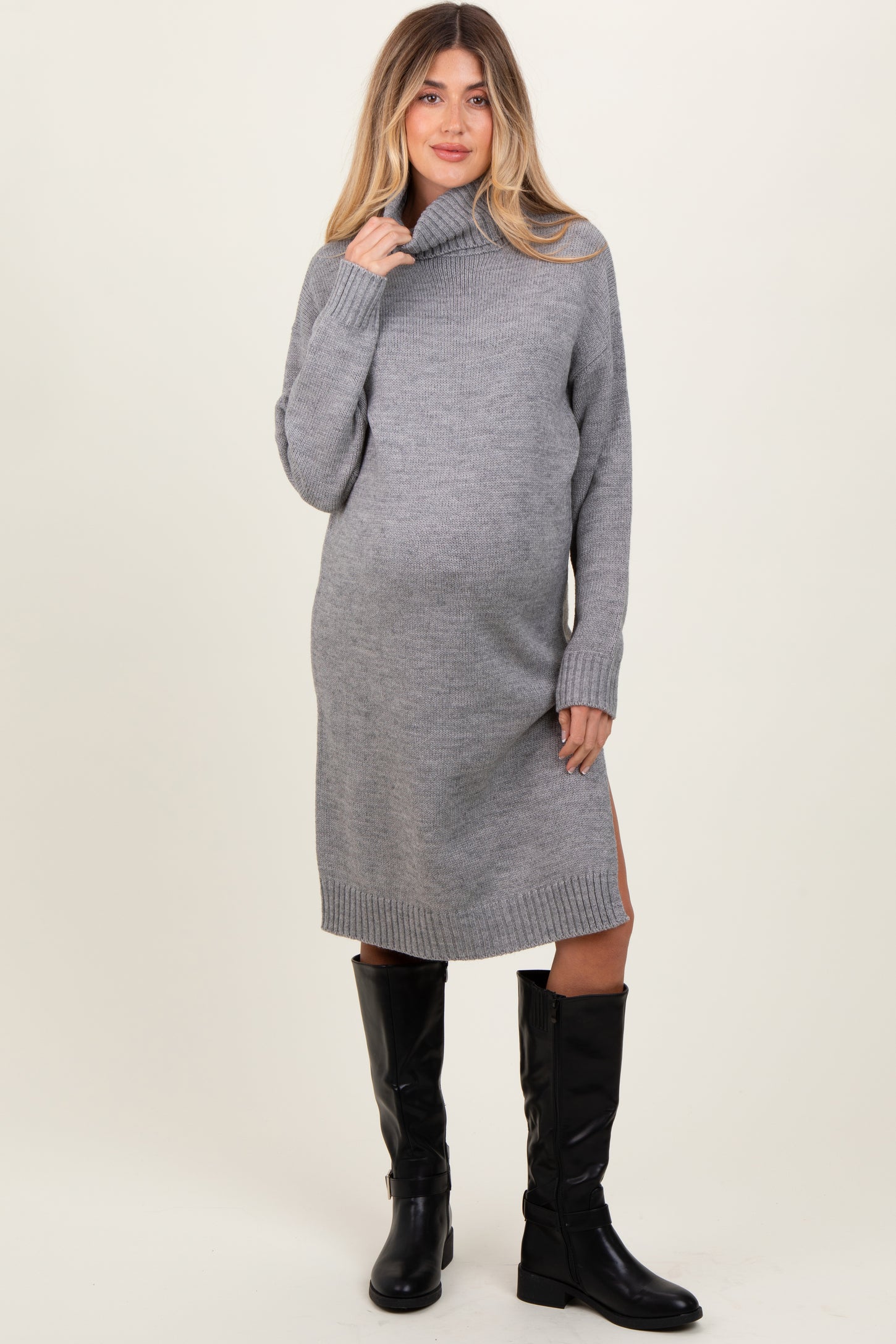 woman wearing a grey turtleneck maternity sweater dress with side slits and knee-high black boots, showcasing her baby bump
