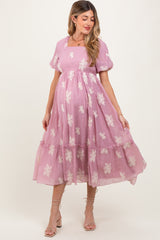 woman wearing a pink maternity dress with floral embroidery and a tiered skirt, showcasing her baby bump