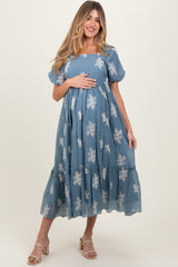 woman wearing a blue floral embroidered maternity dress, holding her baby bump, with puff sleeves and a tiered skirt