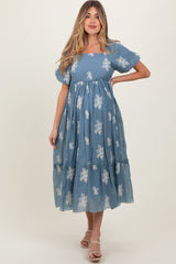 woman wearing a blue maternity dress with white floral embroidery and a tiered hem, smiling and posing with one hand on her hip