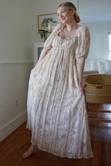 woman wearing a beige floral maxi dress with puff sleeves and a fitted bodice, posing in a bright room