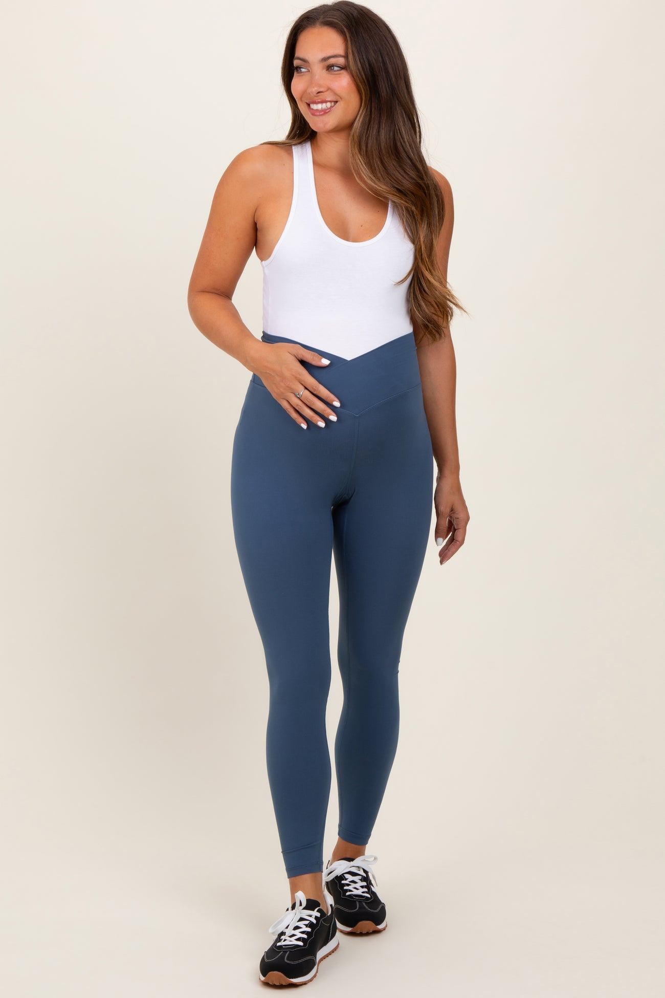 woman wearing a white tank top and blue maternity leggings, holding her baby bump while smiling