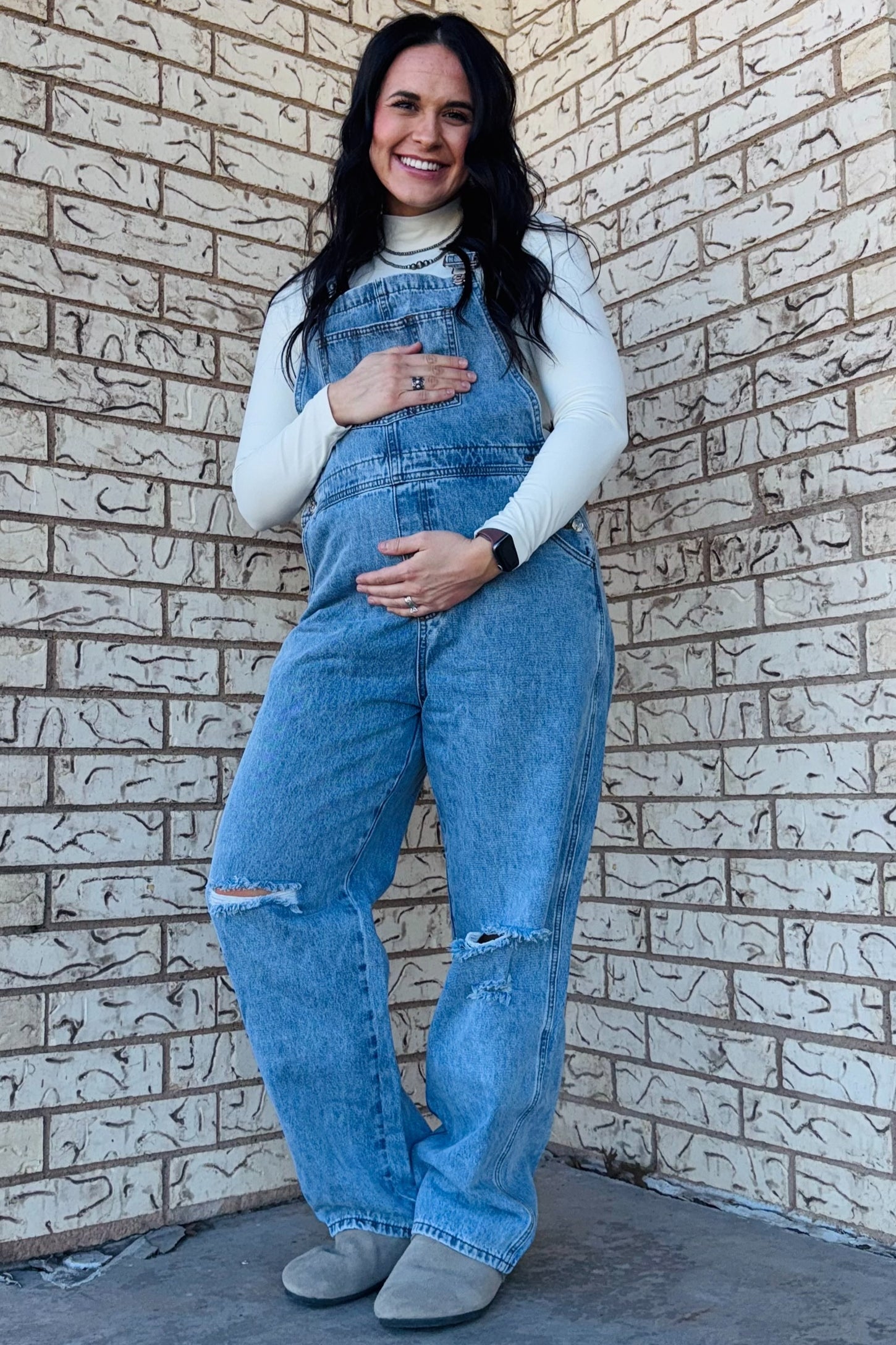 woman wearing a light blue maternity overall with distressed details and holding her baby bump