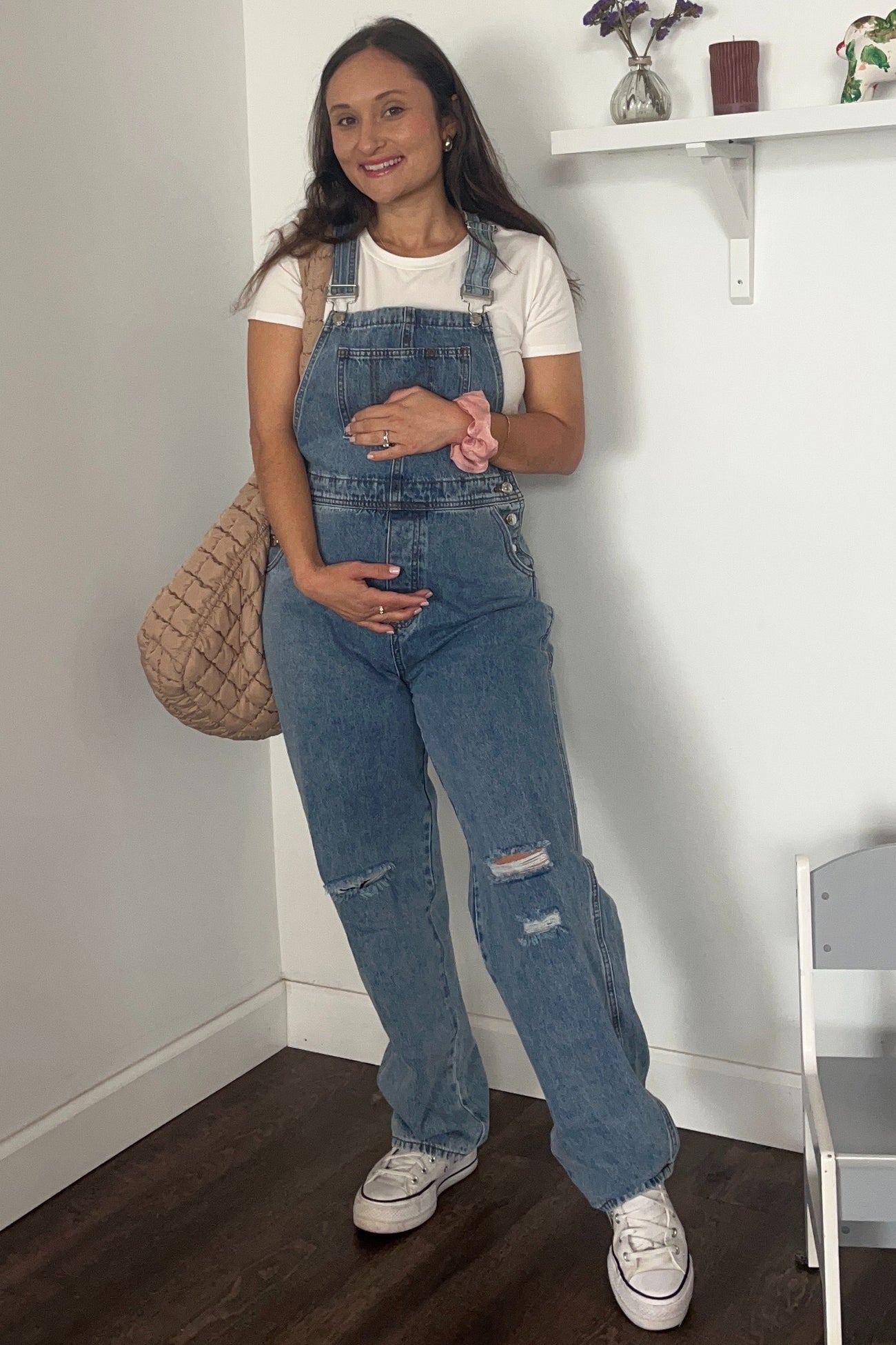 woman wearing a light blue maternity denim overall with rips, holding her baby bump and carrying a beige bag