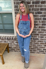woman wearing a pink tank top and denim overalls, holding her baby bump, standing outside against a brick wall