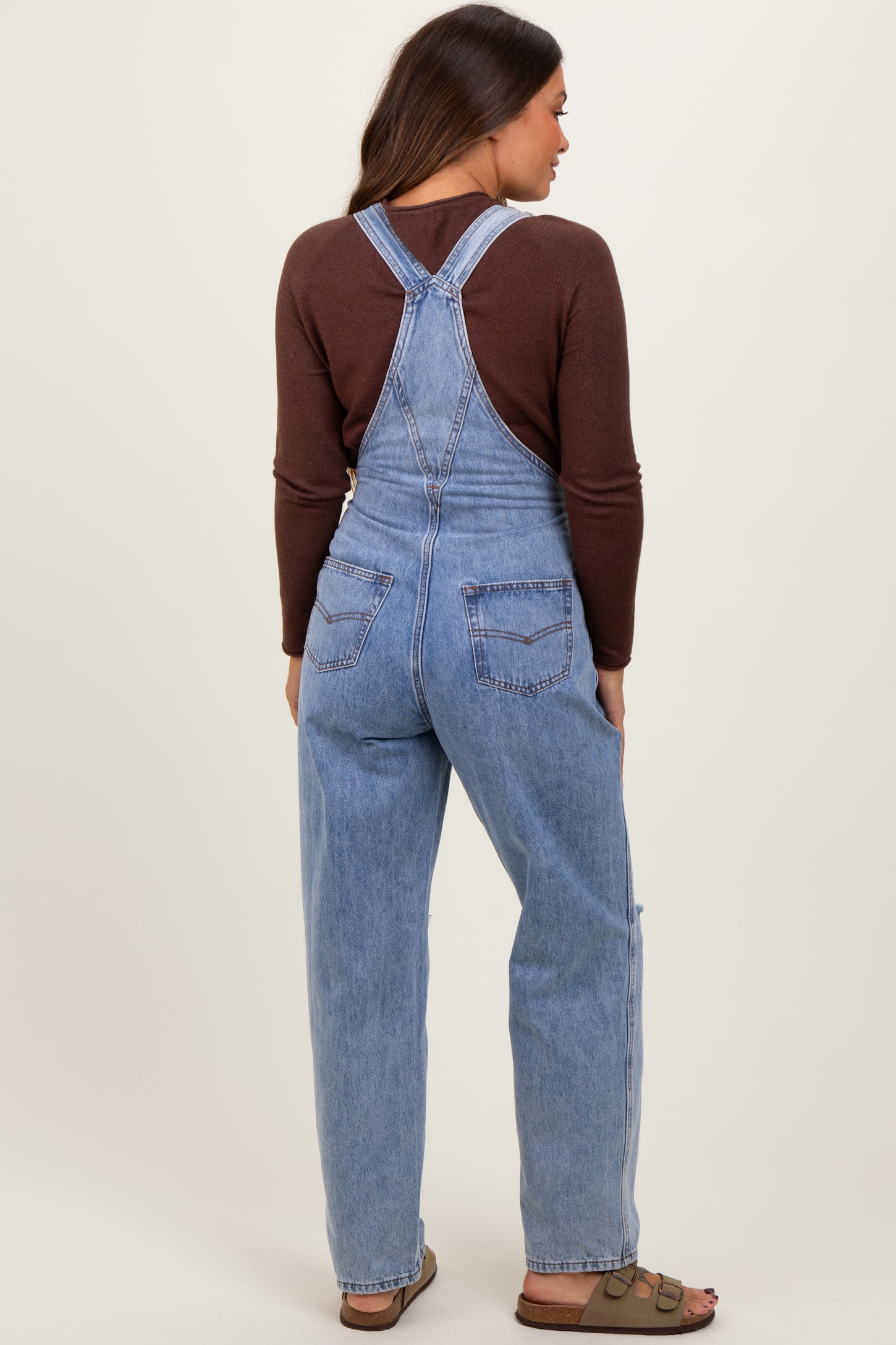 woman wearing a brown long sleeve top and light blue maternity overalls, viewed from the back