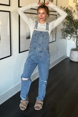woman wearing a light gray long sleeve top and distressed denim overalls, displaying a baby bump