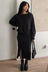 woman wearing a dark gray cropped sweater and matching long skirt with a slit, holding a small black handbag