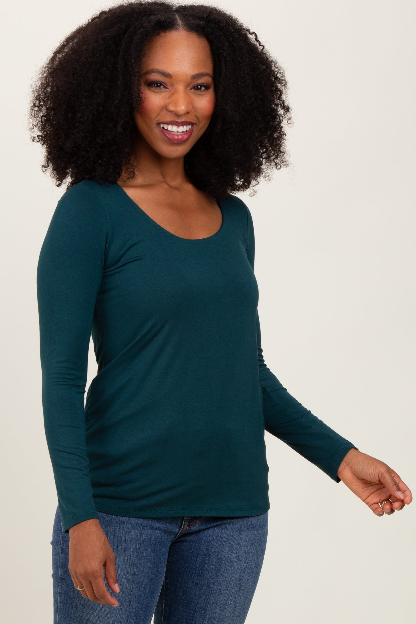 woman wearing a teal long sleeve top and blue jeans, smiling and posing to the side