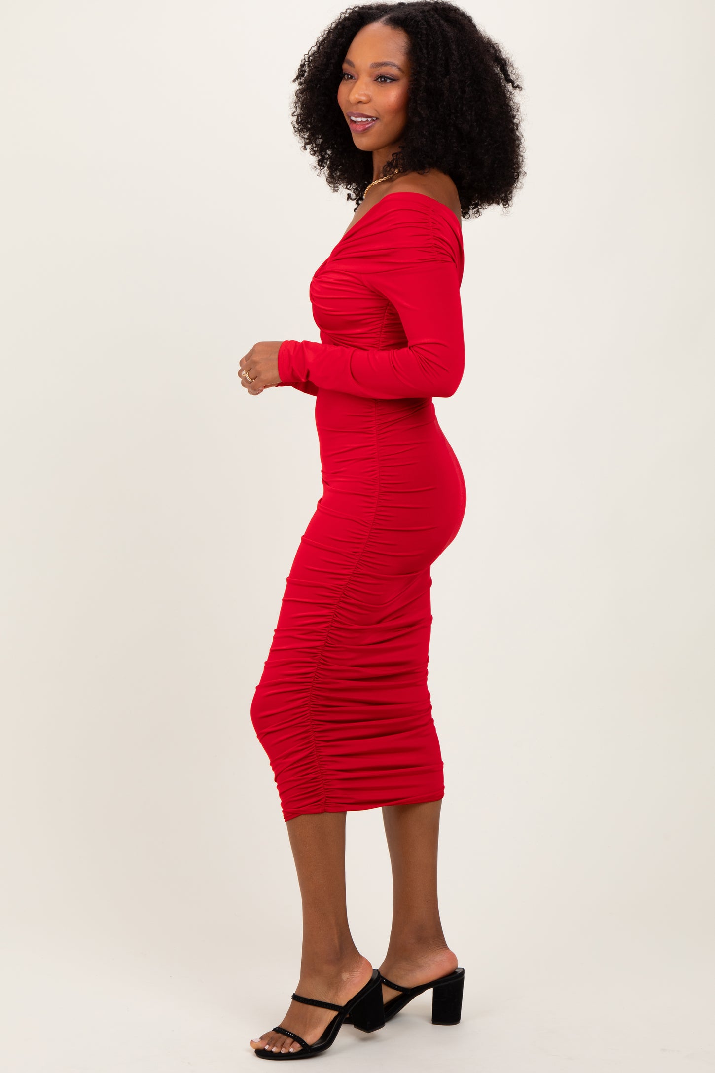 woman wearing a form-fitting red off-the-shoulder midi dress with ruching details and side slit, standing in profile