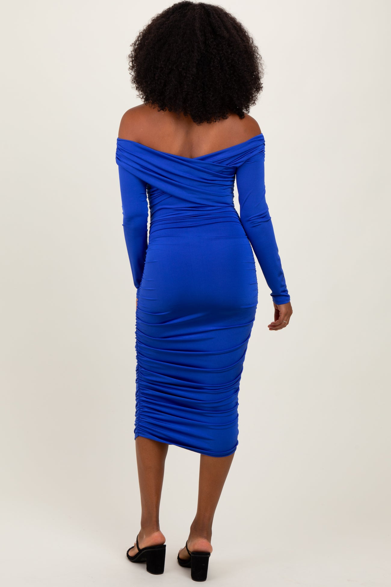 woman wearing a fitted royal blue off-the-shoulder ruched midi dress and black sandals, viewed from the back