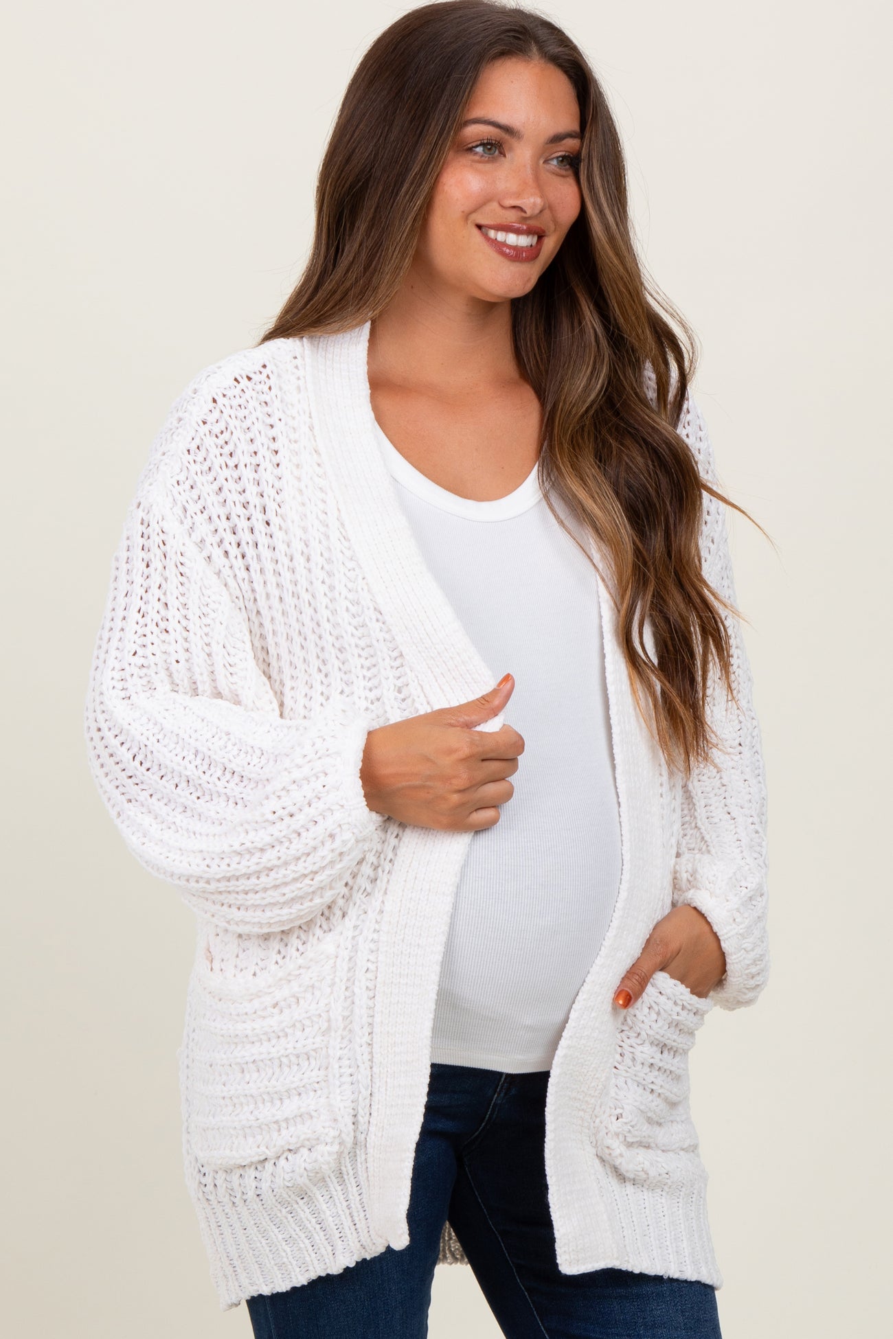 woman wearing a white open-knit maternity cardigan over a white top and showing her baby bump