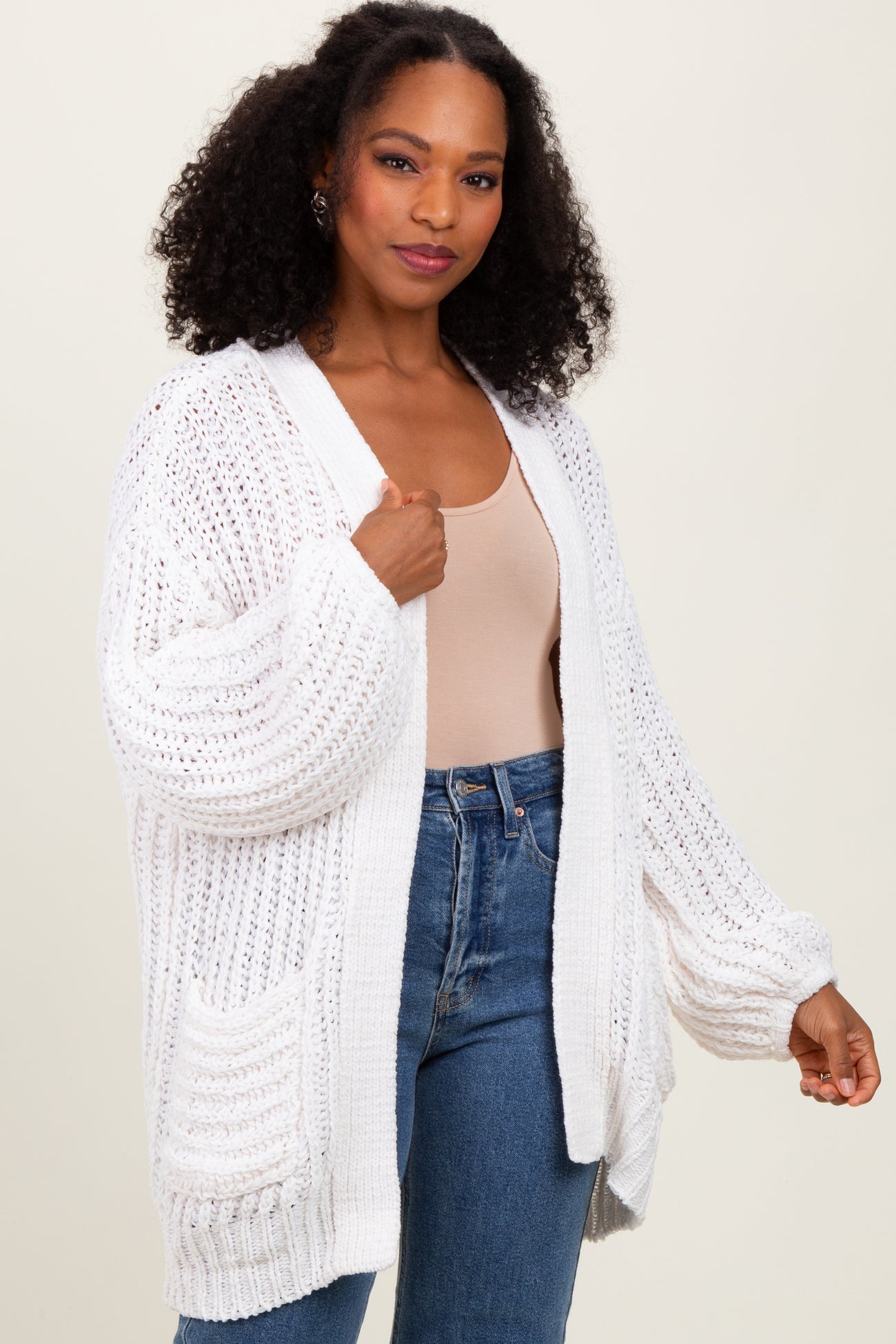 woman wearing a white oversized cardigan with textured knitting and pockets, paired with a beige tank top and denim jeans