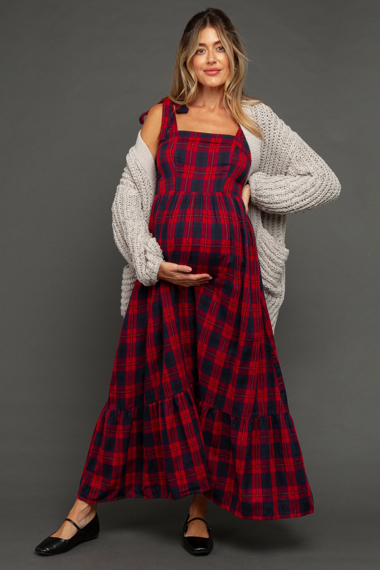 woman wearing a red and navy plaid maternity dress with a ruffled hem and a cozy gray cardigan, holding her baby bump