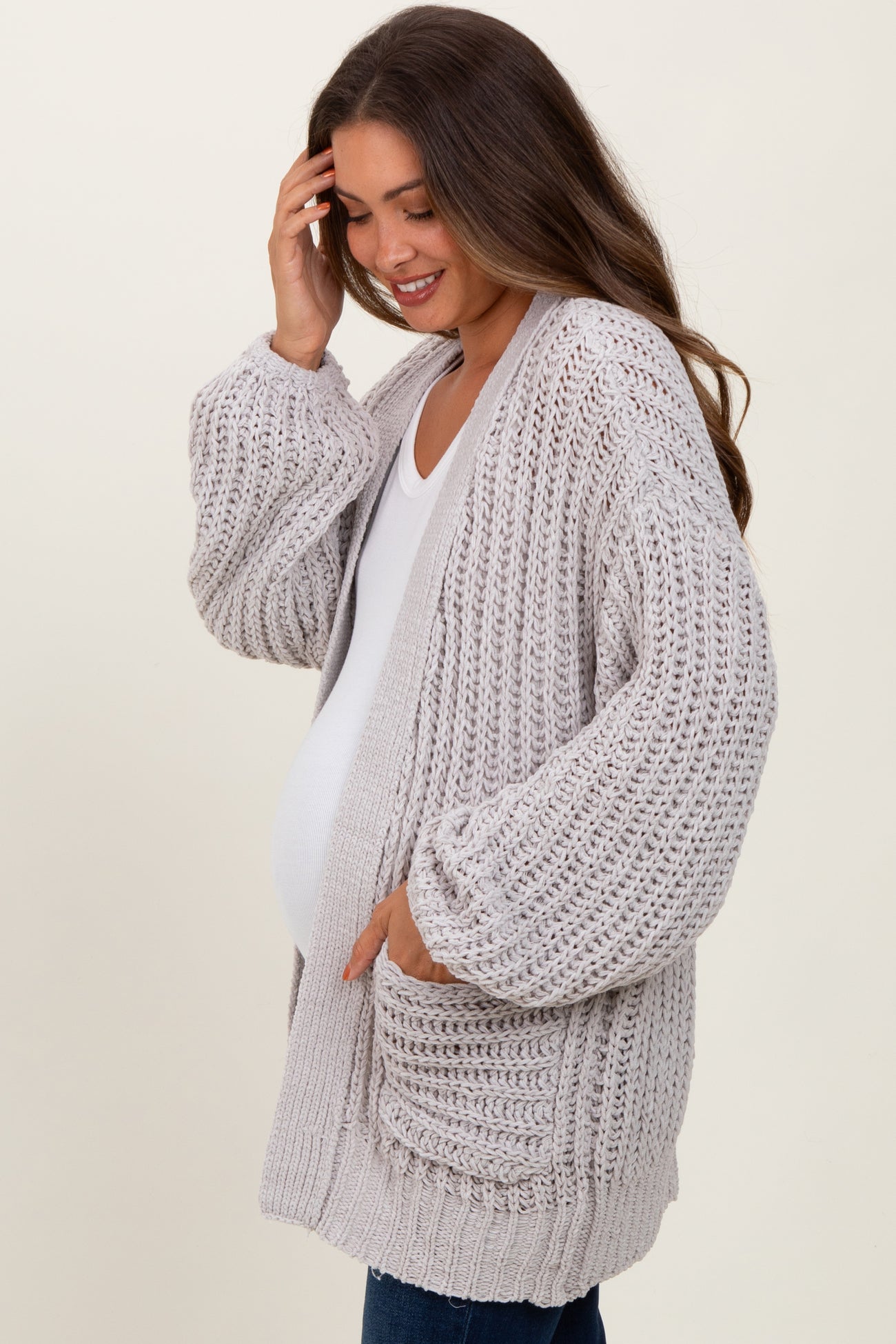 woman wearing a light grey knitted maternity cardigan with pockets, gently touching her hair and showing her baby bump