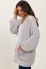 woman wearing a light grey knitted maternity cardigan with pockets, gently touching her hair and showing her baby bump