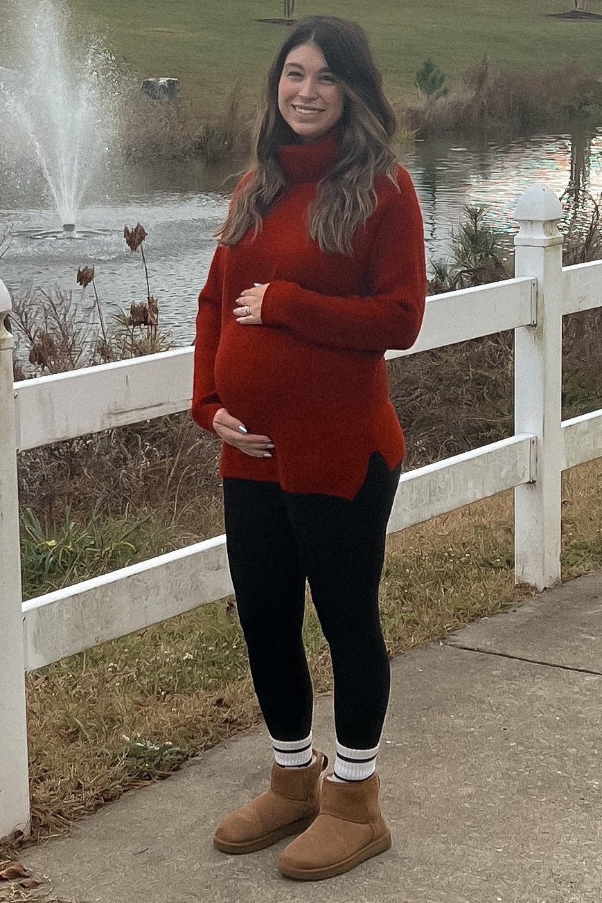 woman wearing a red oversized sweater and black leggings, showing her baby bump while standing outdoors