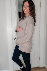 woman wearing a beige oversized maternity sweater and holding her baby bump while standing in front of a door