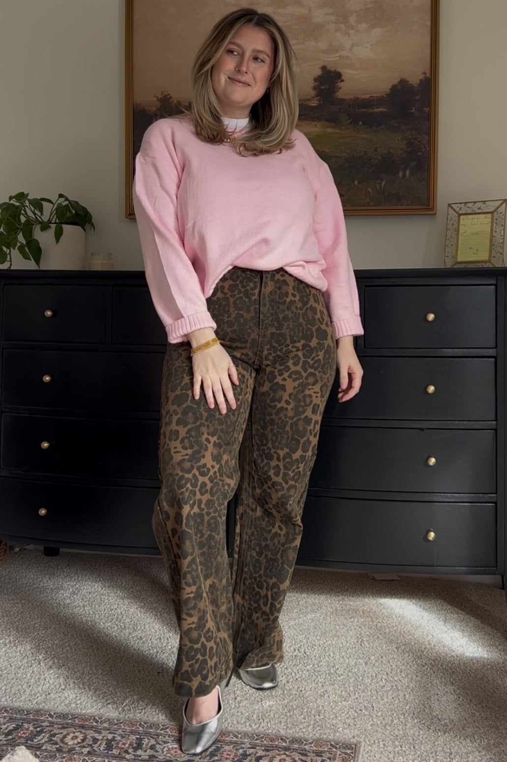 woman wearing a pink sweater and leopard print pants, standing in front of a dresser and smiling