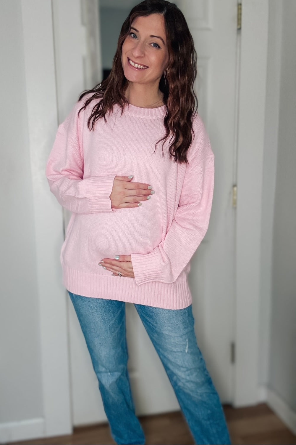 woman wearing a light pink maternity sweater and blue jeans, gently cradling her baby bump