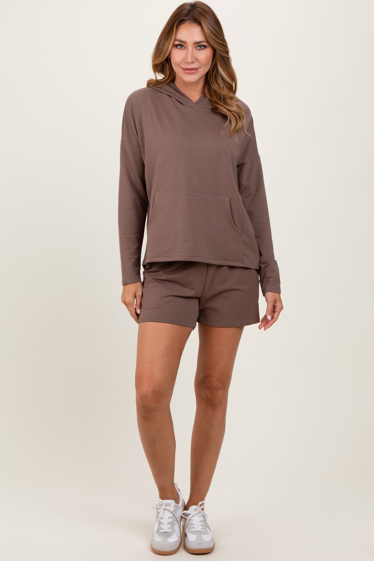 woman wearing a brown hooded sweatshirt with a front pocket and matching brown shorts, styled with white sneakers