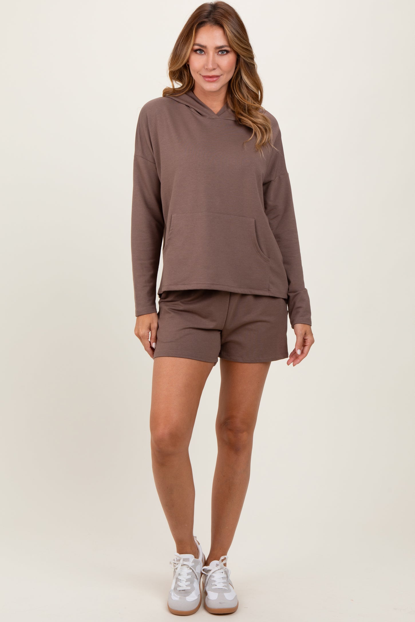 woman wearing a brown hooded sweatshirt with a front pocket and matching brown shorts, styled with white sneakers