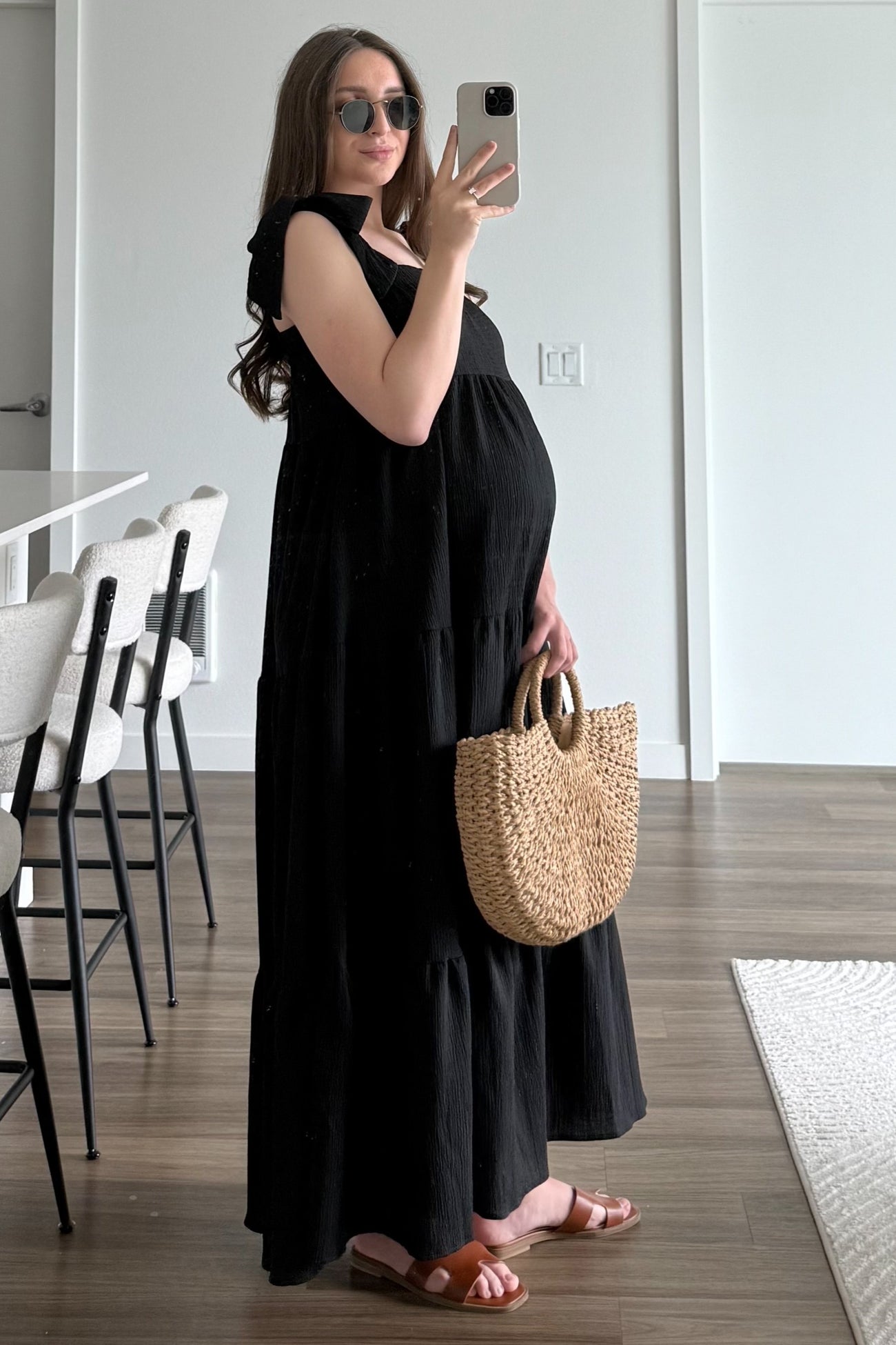 woman wearing a black tiered maternity dress, holding a straw bag, and showing off her baby bump in a bright room